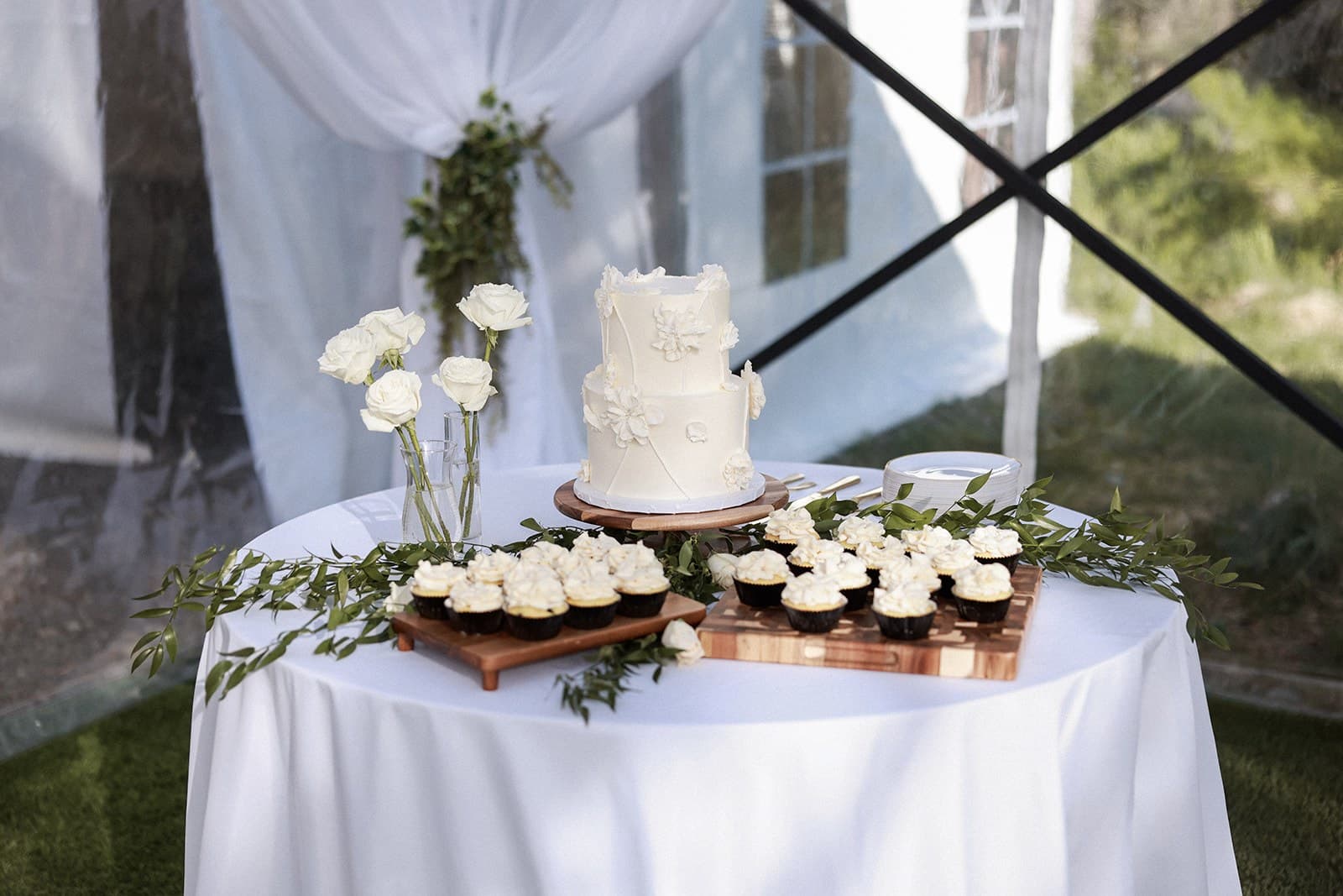 White wedding cake on a draped table with white roses and cupcakes