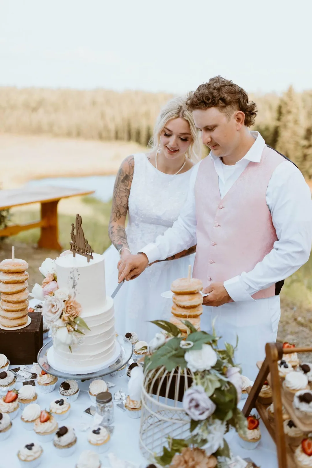 Couple cutting their wedding cake lakeside with a donut tower