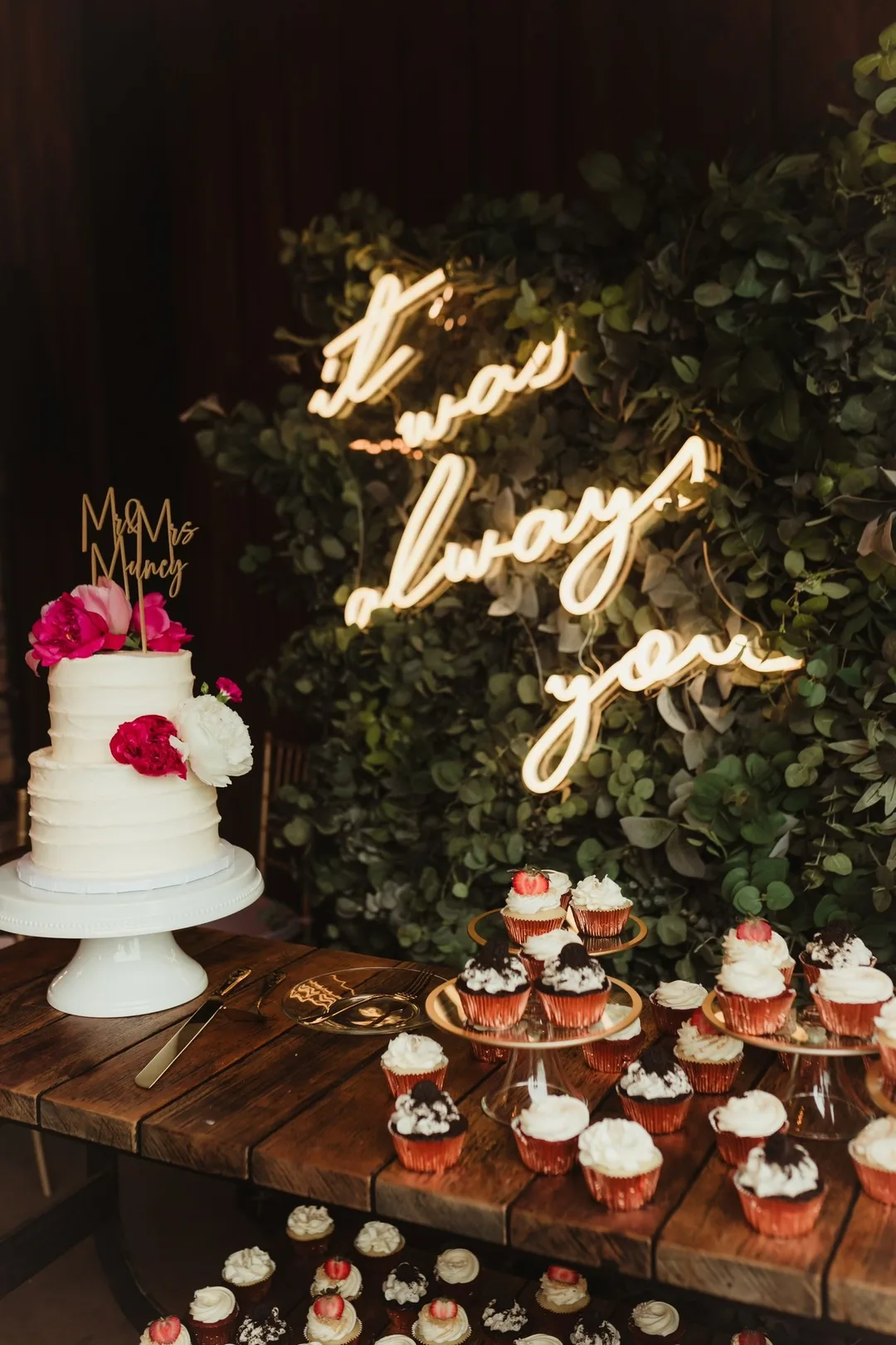 White ruffle wedding cake with pink florals under a neon sign, cupcakes below